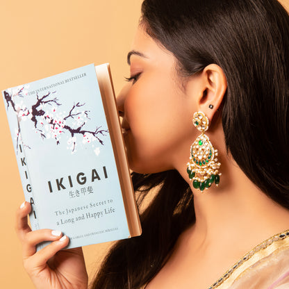 A person wearing green and gold traditional earrings, kissing a book titled 'IKIGAI'.