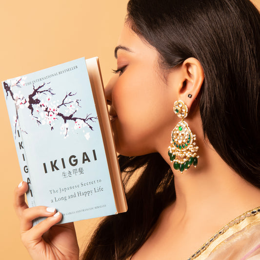 A person wearing green and gold traditional earrings, kissing a book titled 'IKIGAI'.